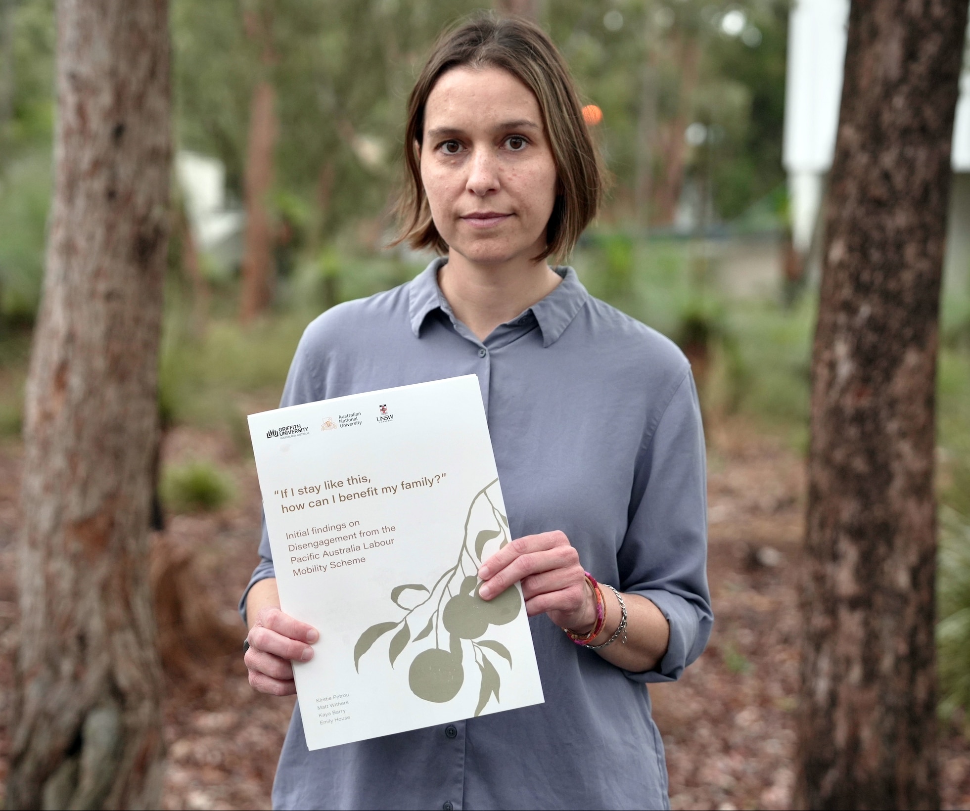 A woman wearing a grey button up shirt stands between trees in a holding a printed booklet. She has a short cropped hair style.