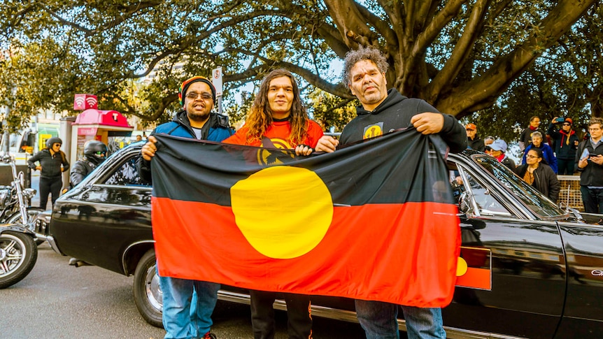 Three people hold up Aborginal flag under a large tree.