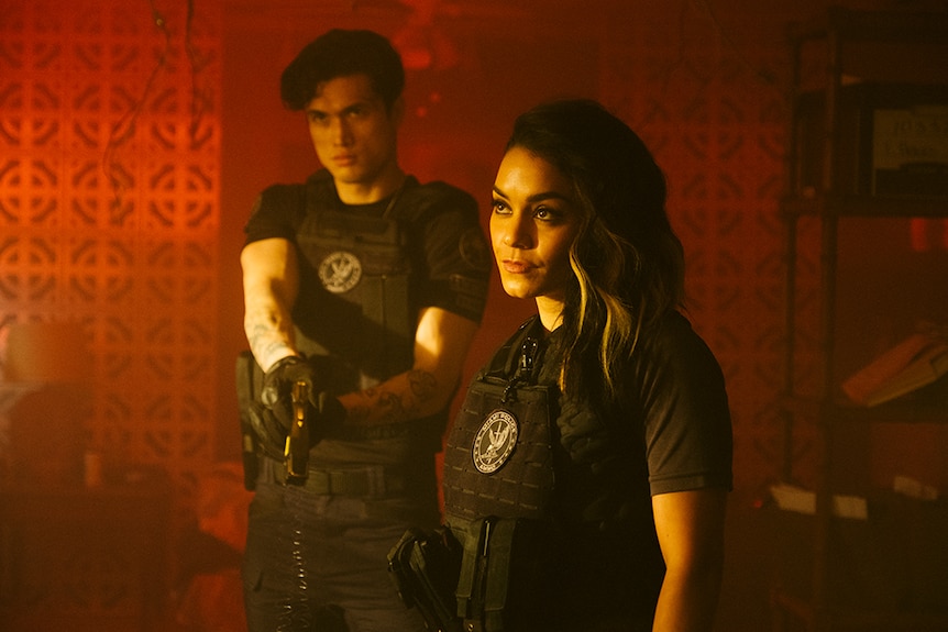 A man and woman dressed in bullet proof vests are warmly illuminated in red hued and hazy room with patterned concrete walls.