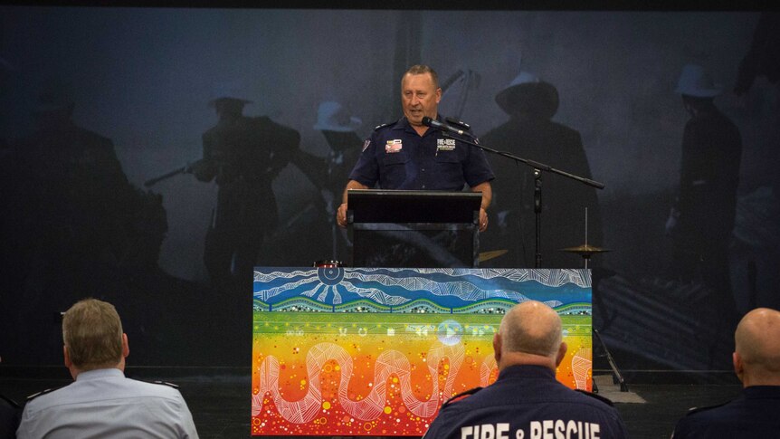 A fireman stands at a podium  behind an indigenous artwork