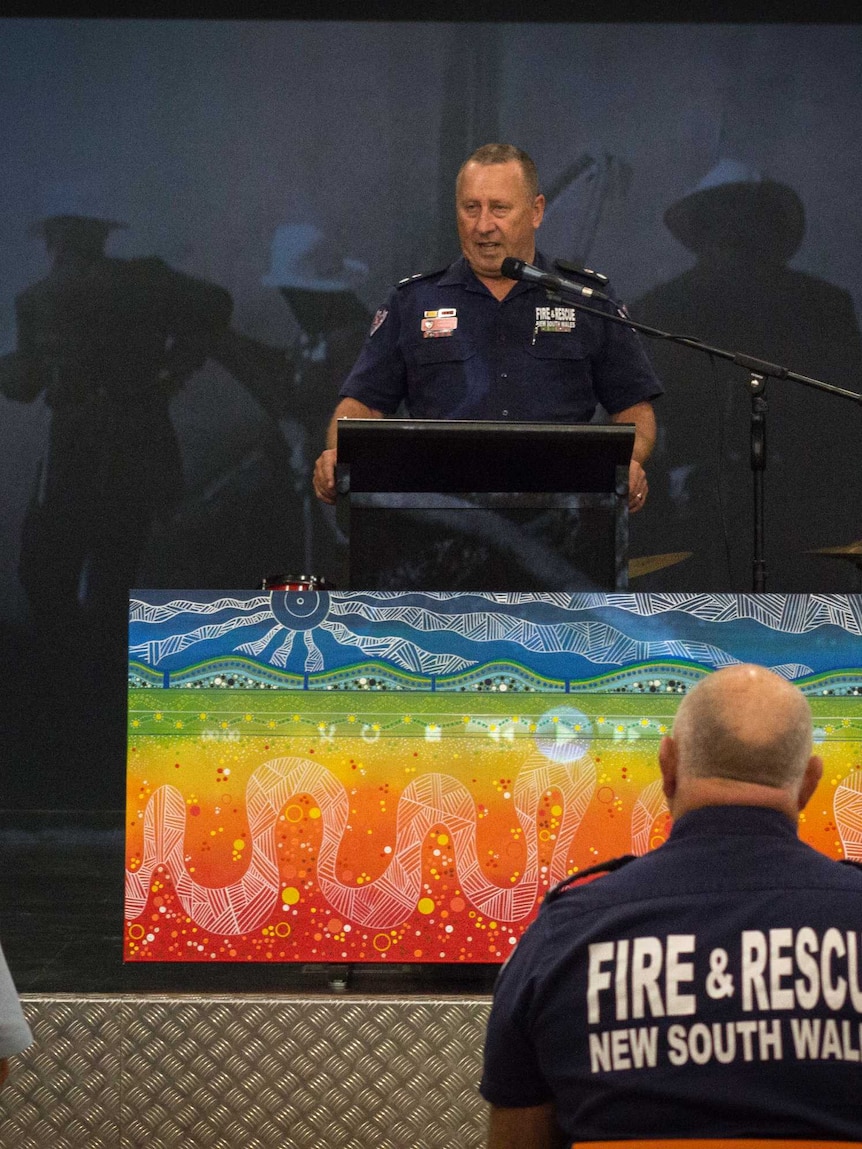 A fireman stands at a podium  behind an indigenous artwork