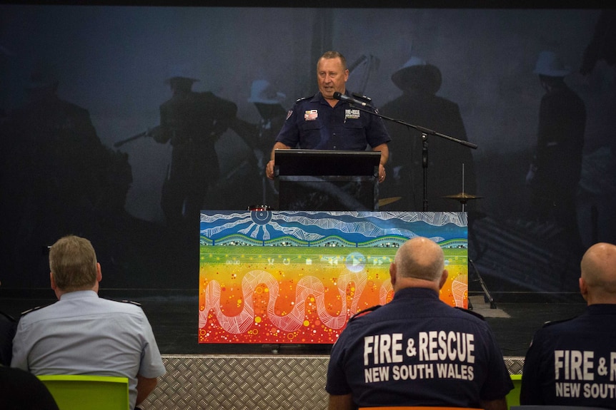 A fireman stands at a podium  behind an indigenous artwork