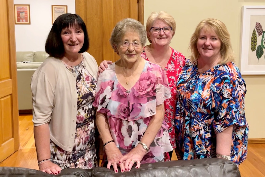 An elderly woman in a floral dress surrounded by three middle-aged women smile for the camera.  