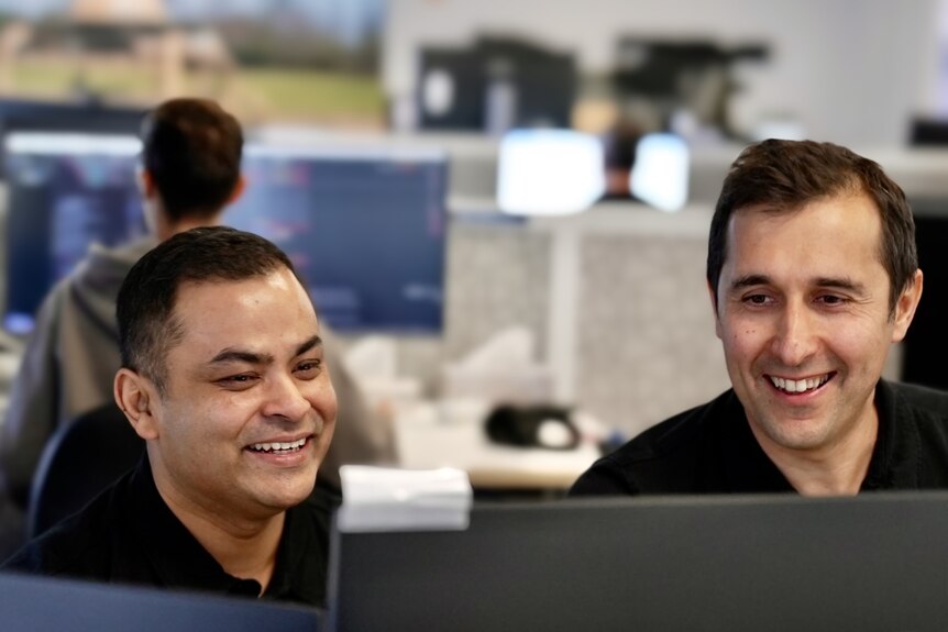 Deux hommes, tous deux en polos noirs, sourient tous les deux, regardant un ordinateur.