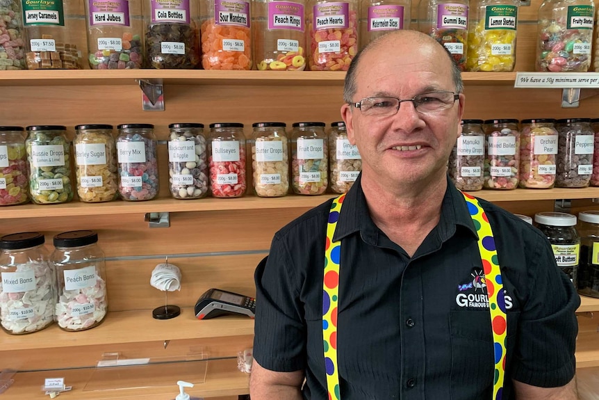 An older man wearing glasses stands in front of lolly jars on a shelves.