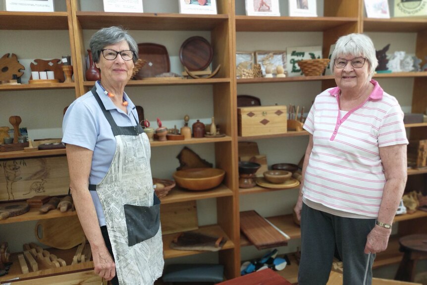 Two women stand in front of a shelf filled with items which have been worked on by their group. 