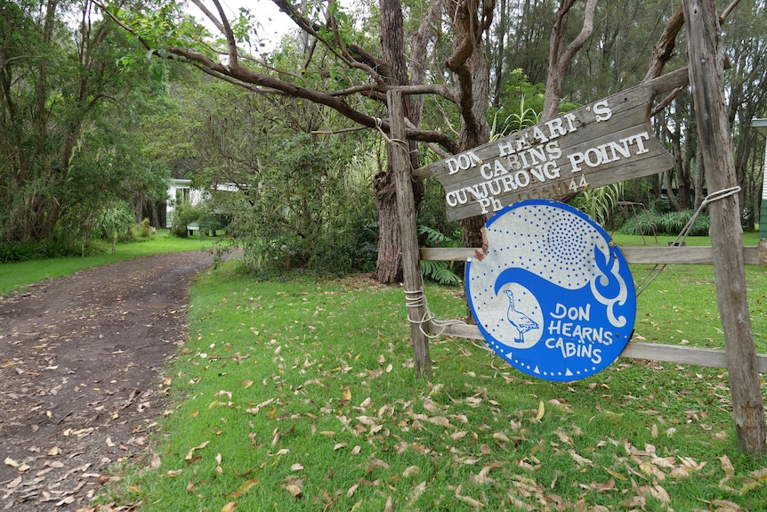 Wooden sign with blue and white paint standing out the front of a tree-lined driveway and green lawn.