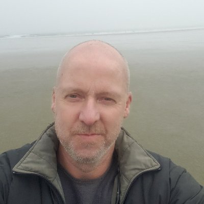 A bald man in a jacket smiling near the ocean