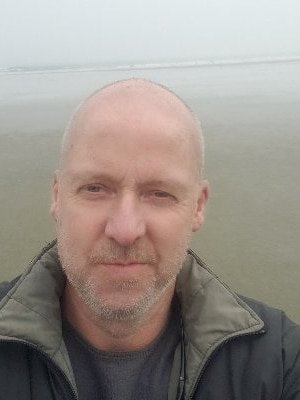 A bald man in a jacket smiling near the ocean