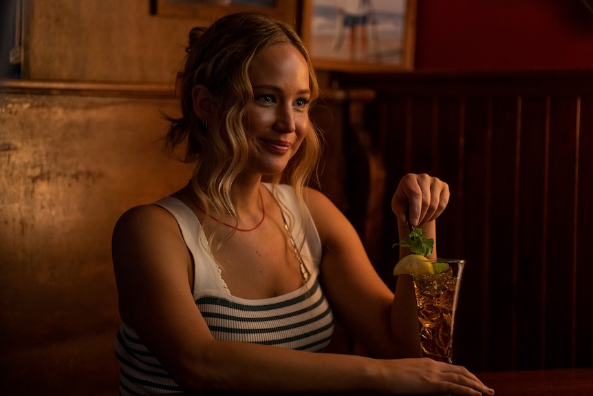 A blonde white woman wearing a white and black striped singlet sits in a warmly-lit bar swilling a cocktail.