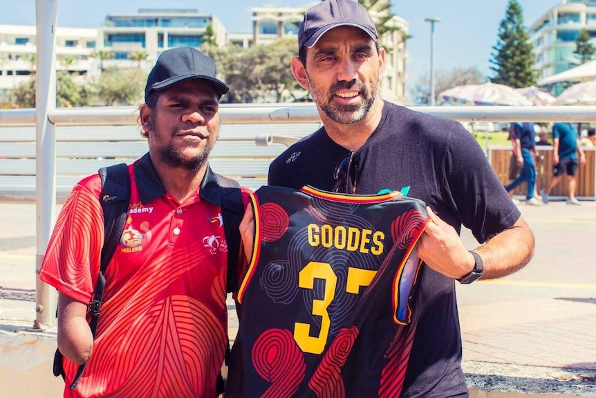 A young man with half his arm missing poses with footballer Adam Goodes, who holds up a shirt that says 'Goodes'.