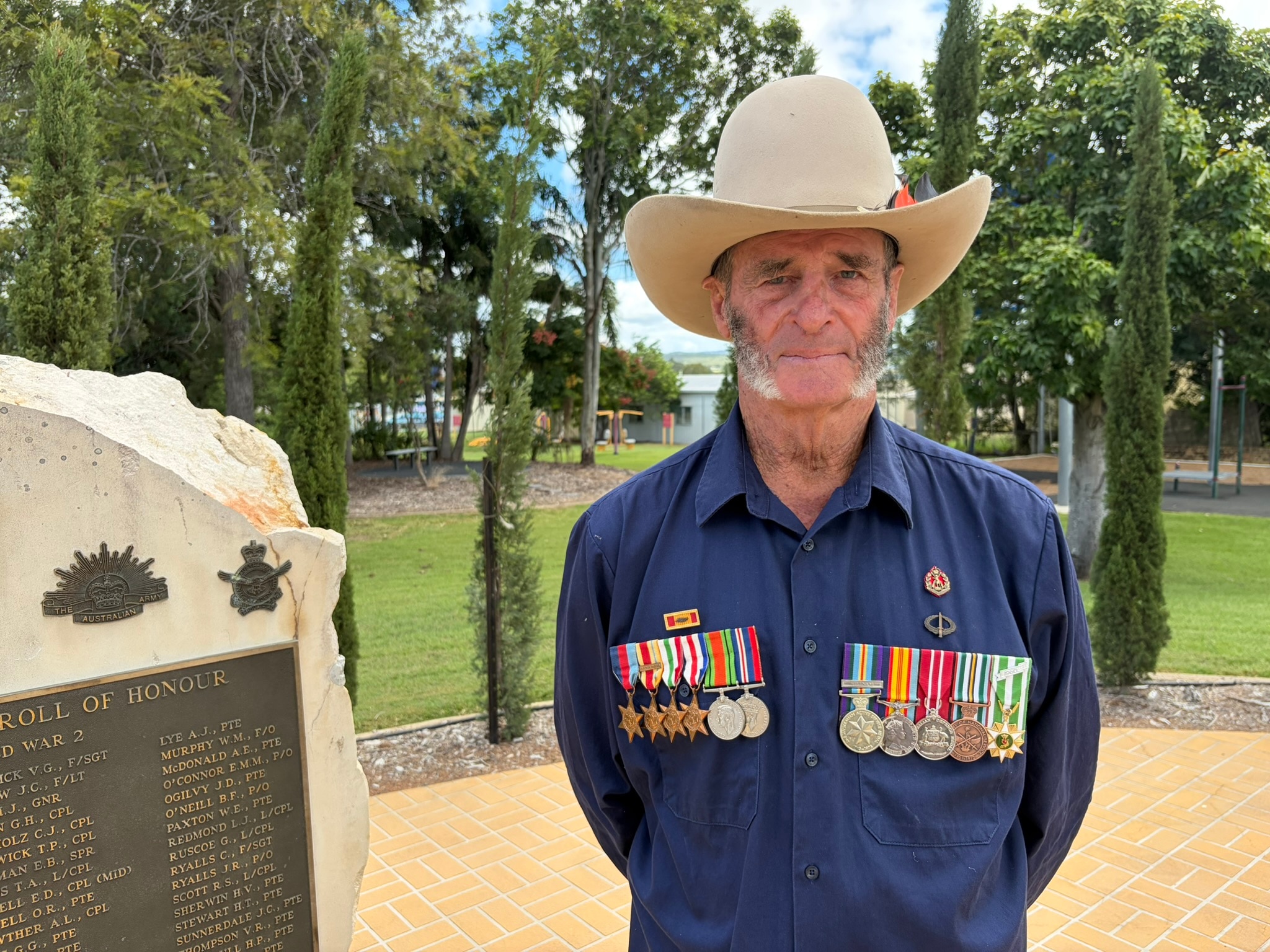 Un hombre con un gran sombrero y una hilera de medallas a cada lado del pecho se encuentra junto a un monumento a los caídos.