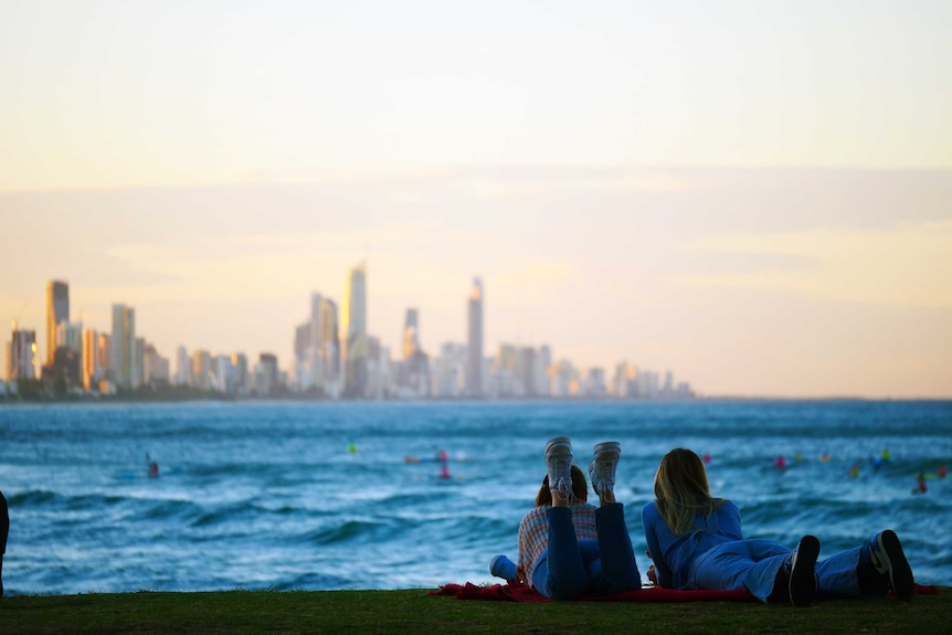 Two young women lie on their stomachs on a blanket on a grassy hill looking out at water, to a highrise peninsula in background.