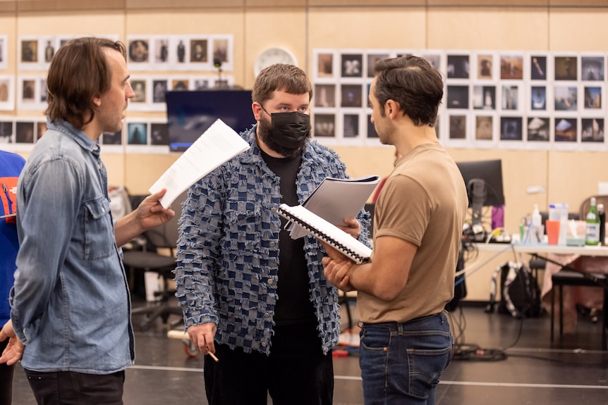 Three men, aged from their mid-30s to early 40s, stand near each other, each holding scripts in a rehearsal space