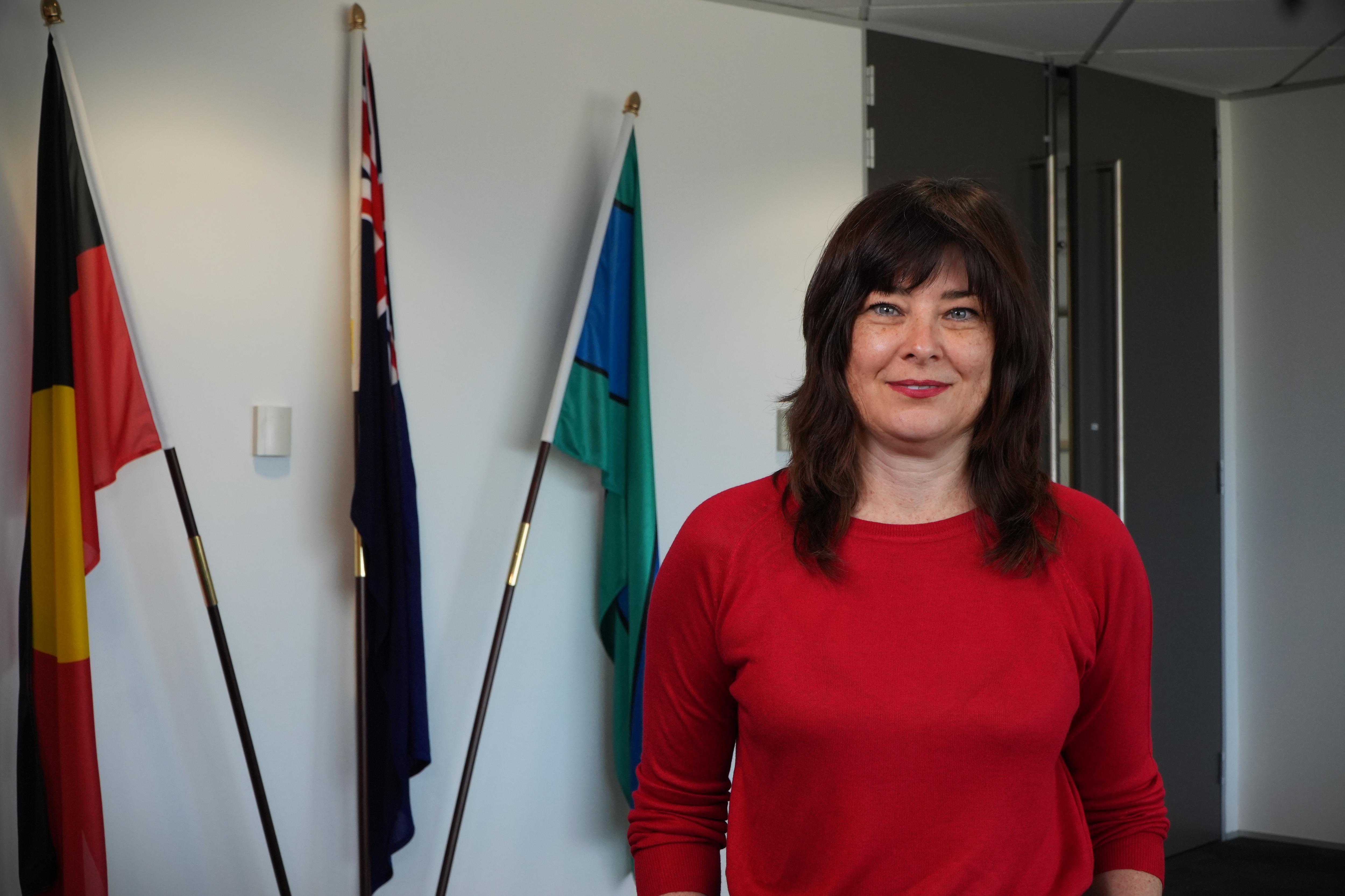 Una mujer sonriente que llevaba una camisa roja brillante con tres mástiles detrás de ella en un ambiente de oficina.