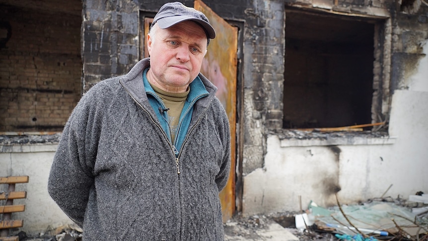 A man wearing a cap and jumper stands in front of his house and a destroyed car.