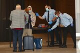 2 travellers stand before a Perth hotel desk. 2 masked security guards offer to take their luggage.