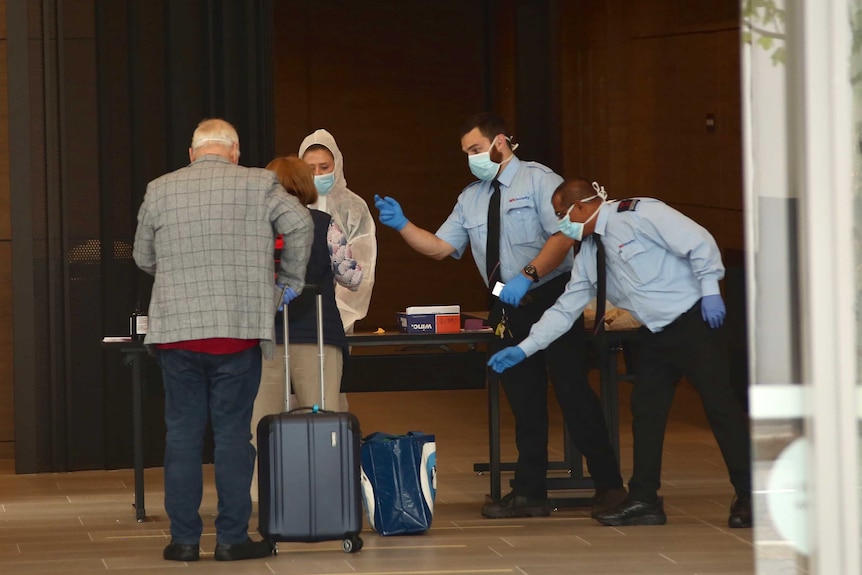 2 travellers stand before a Perth hotel desk. 2 masked security guards offer to take their luggage.