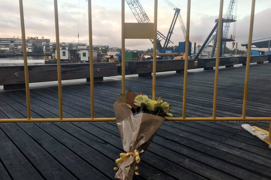 A bunch of flowers lean against a fence, with a crane barge visible behind.