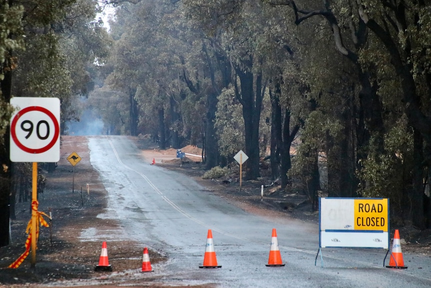 "Road closed" and speed limit signs beside a rural road, with burnt bushland beyond.