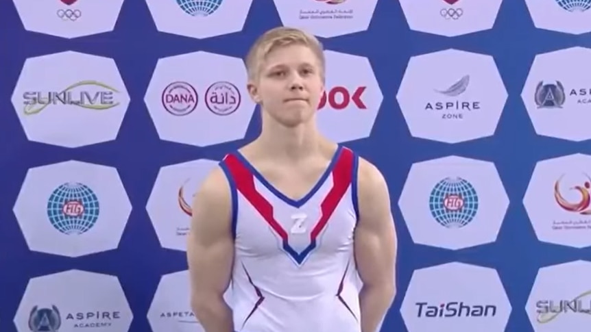 A gymnast stands on the third-place dais of a podium with a Z symbol on his chest