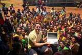 A man holds up a laptop with a sea of children behind him.