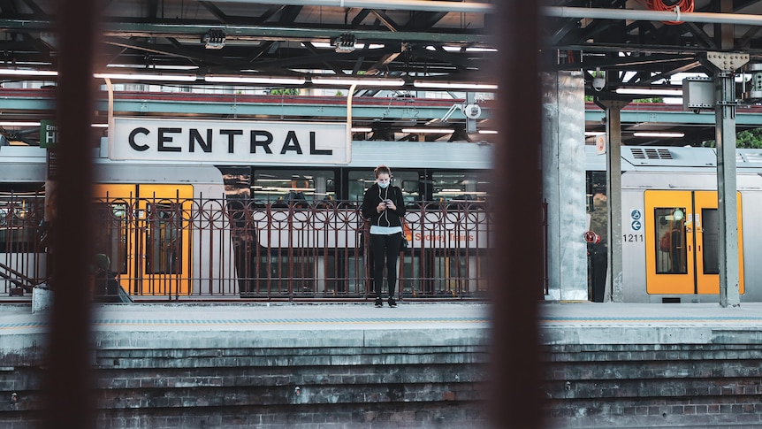 Una mujer enmascarada revisando su teléfono mientras está de pie en una plataforma de tren