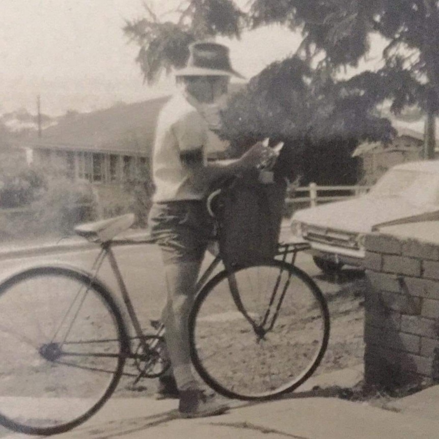 a man stands holding up his push bike between his legs sorting letters from his postie bag. He is wearing shorts, a tshirt n hat