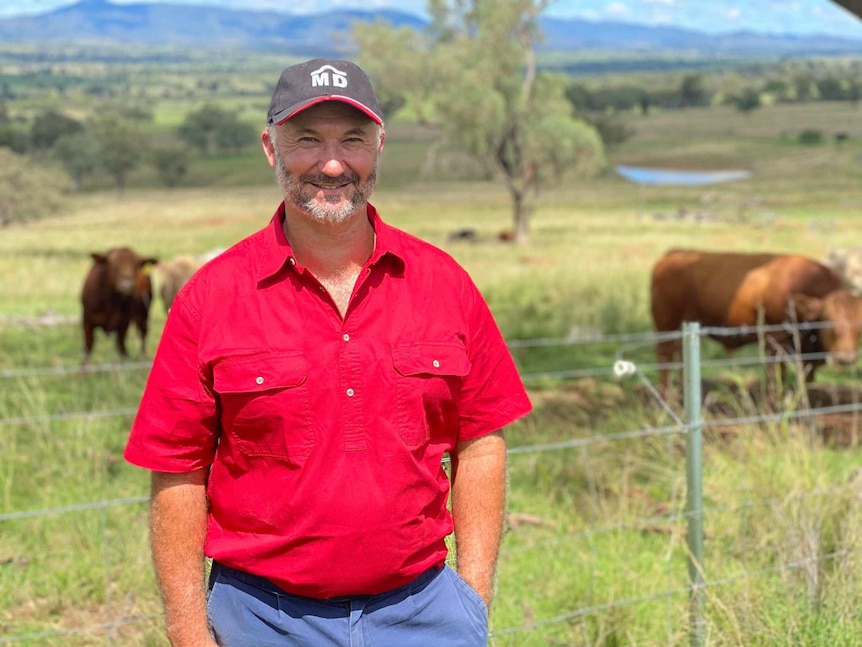 A man in a red shirt smiles at the camera as he stands in front of a green paddock with cattle grazing.