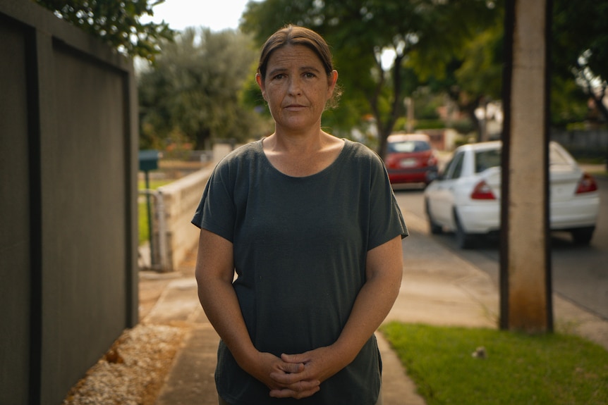 A woman in a dark t-shirt with her hair tied back faces the camera as she stands on a suburban street.