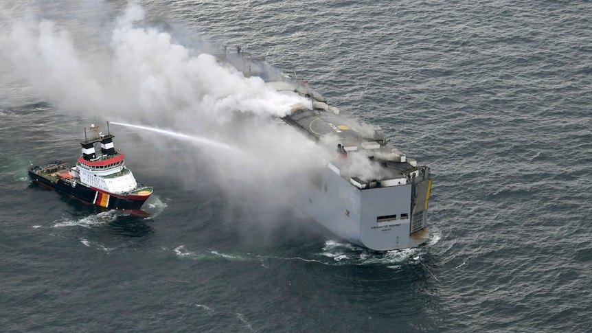 Smoke rises from a burning cargo ship as a nearby boat hoses the fire