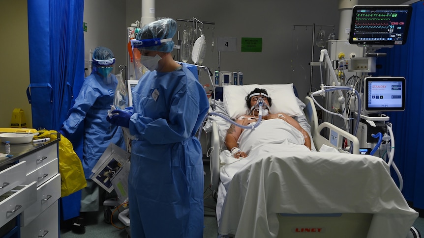 A man lying in bed, while a staff member in PPE walks nearby 