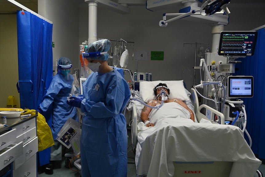 A man lying in bed, while a staff member in PPE walks nearby 