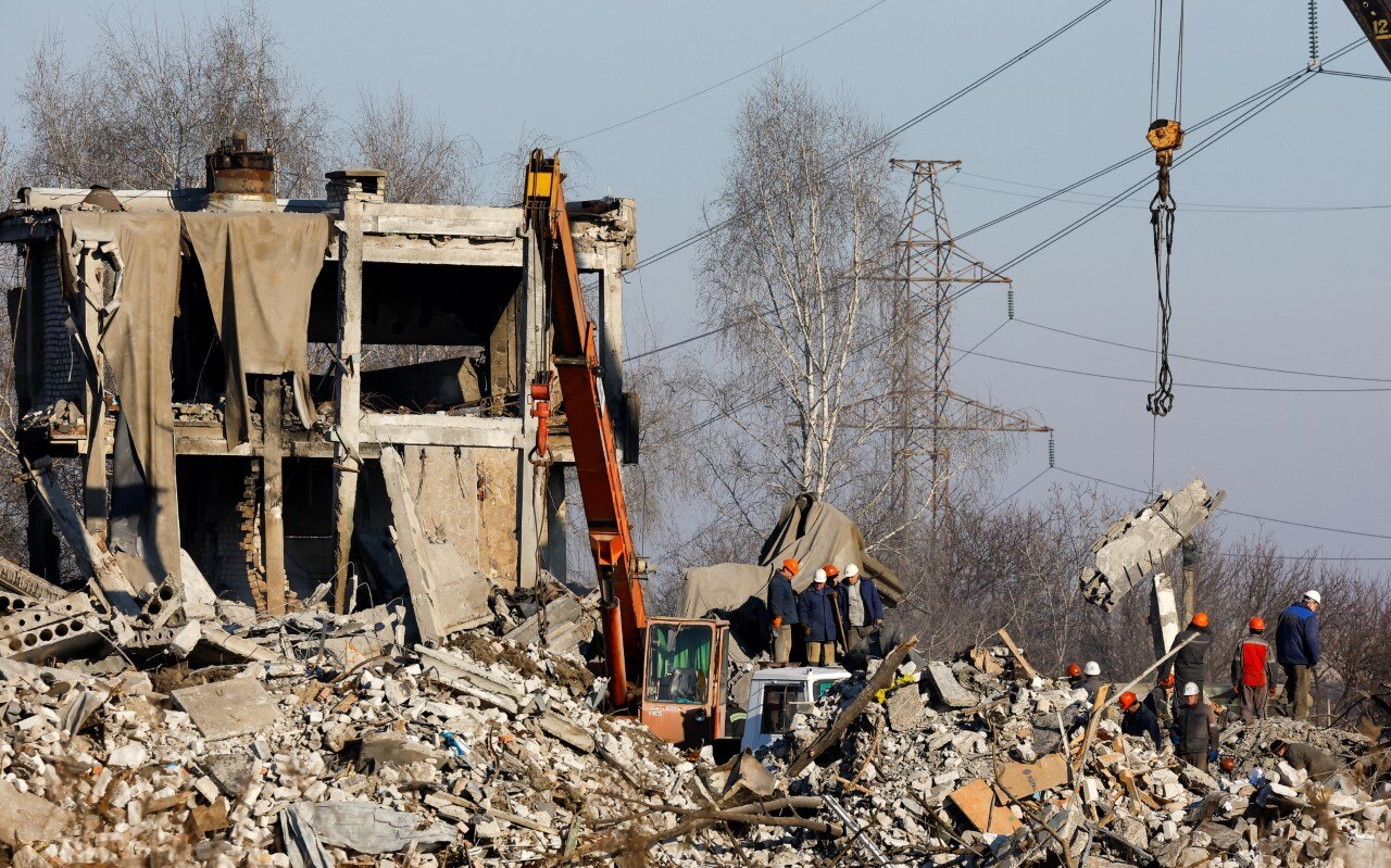 A multi-storey building lies in tatters. Using a crane, workers sift through the rubble