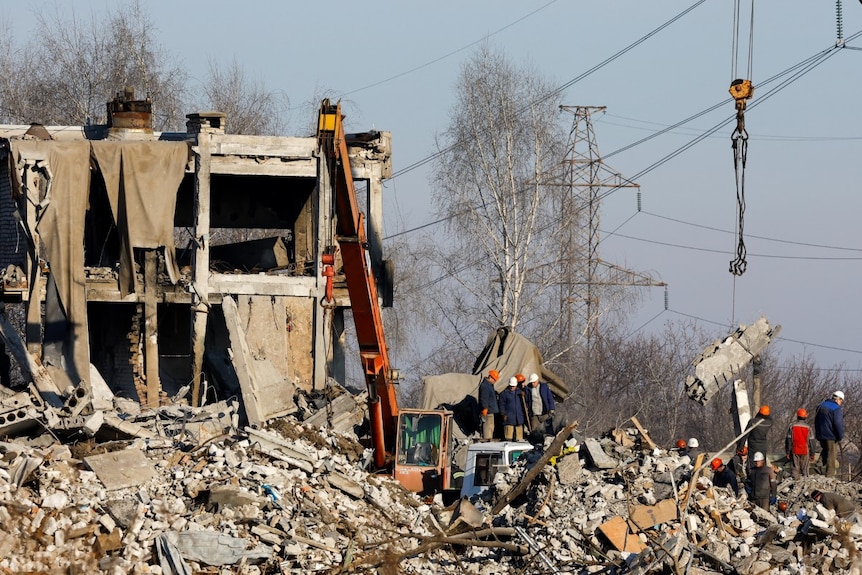 A multi-storey building lies in tatters. Using a crane, workers sift through the rubble