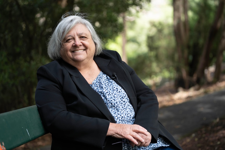 An older woman sits on a park bench, smiling. She has short, grey hair and is wearing a blue, patterned dress and dark jacket.