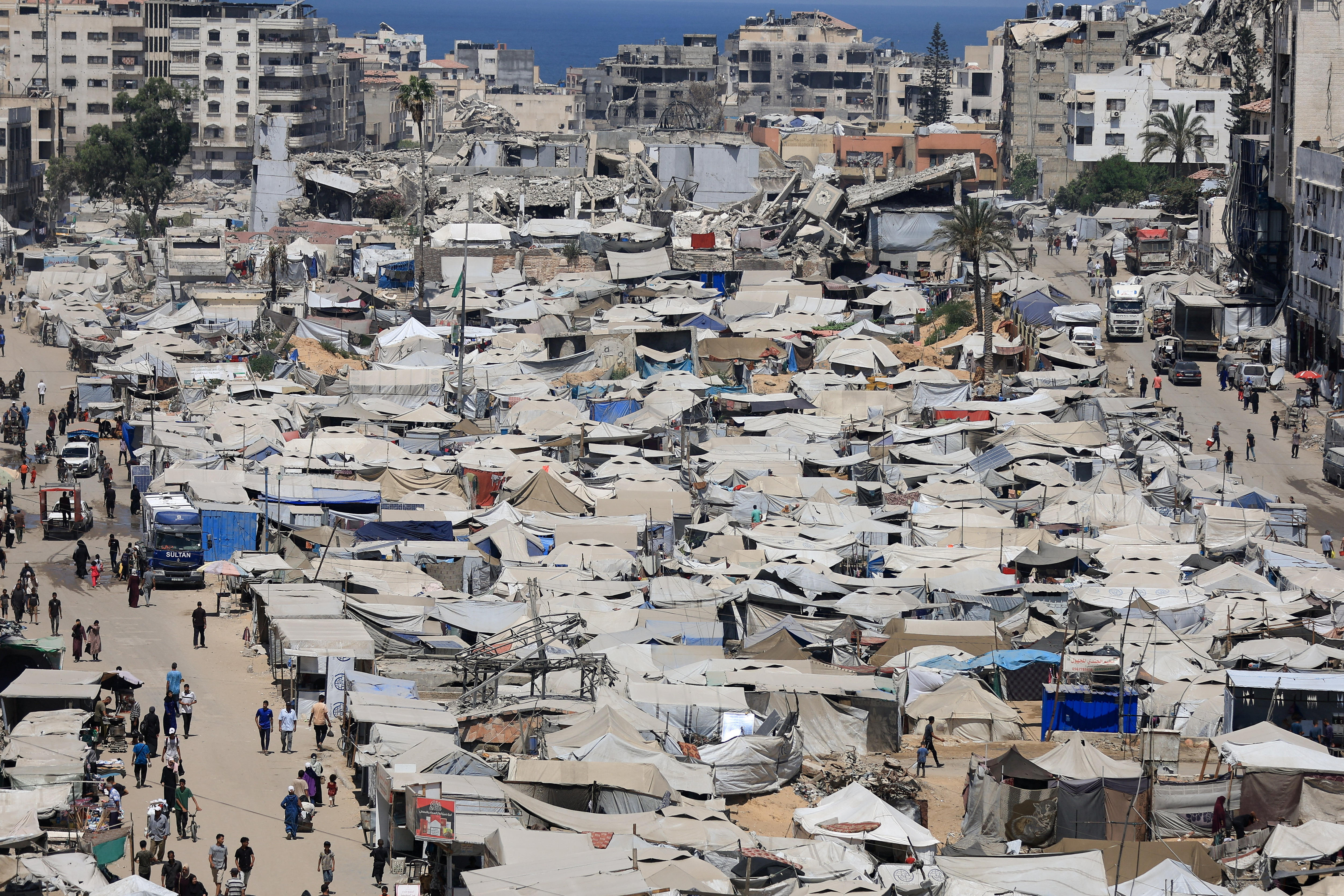 A large dirt area with dozens of make shift tents and people surrounded by destroyed buildings