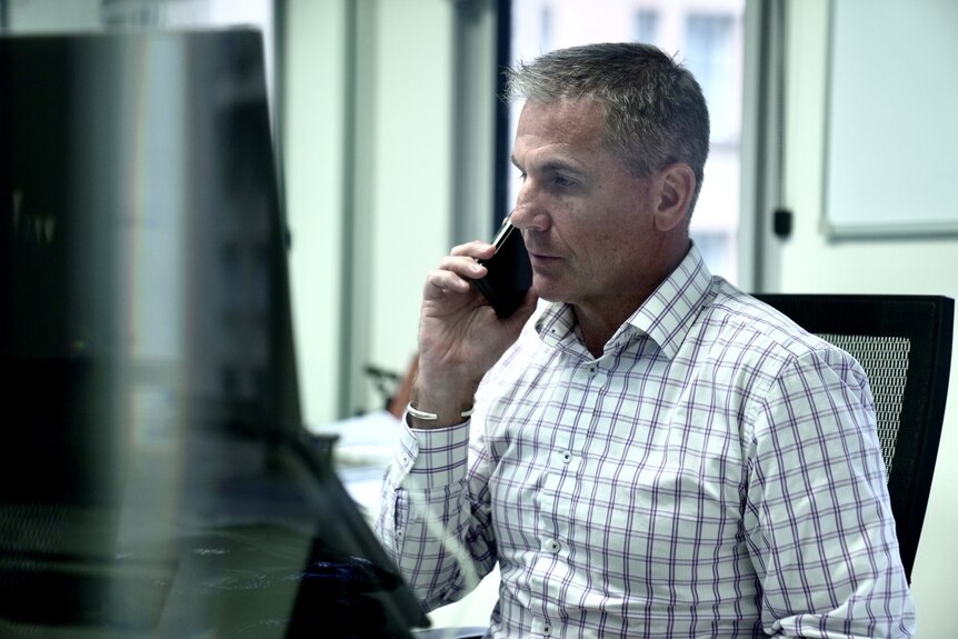 A grey-haired man on a mobile phone as he sits in front of a computer. He's wearing a black and white checked shirt