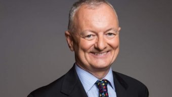 Antony Green smiles. He is wearing a suit and standing against a grey background.