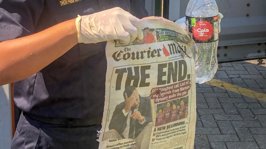 A man in a mask holds up a copy of the Courier Mail newspaper