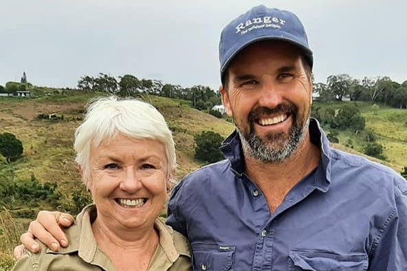 A white haired lady and a bearded man in a cap stand in a hilly field.