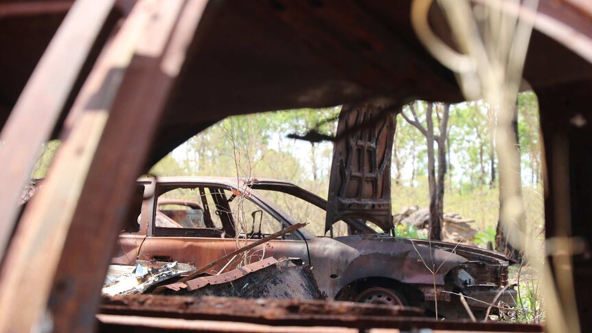 A rusty car can be seen through the open window of another rusty car. Many items are abandoned in the bush.