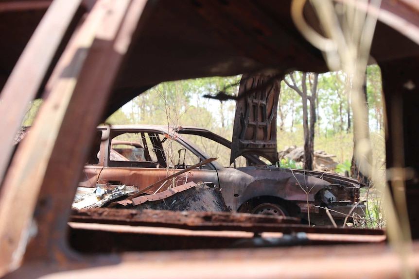 A rusty car can be seen through the open window of another rusty car. Many items are abandoned in the bush.