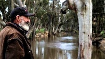 A man with a grey beard looks over a tree-lined body of water 