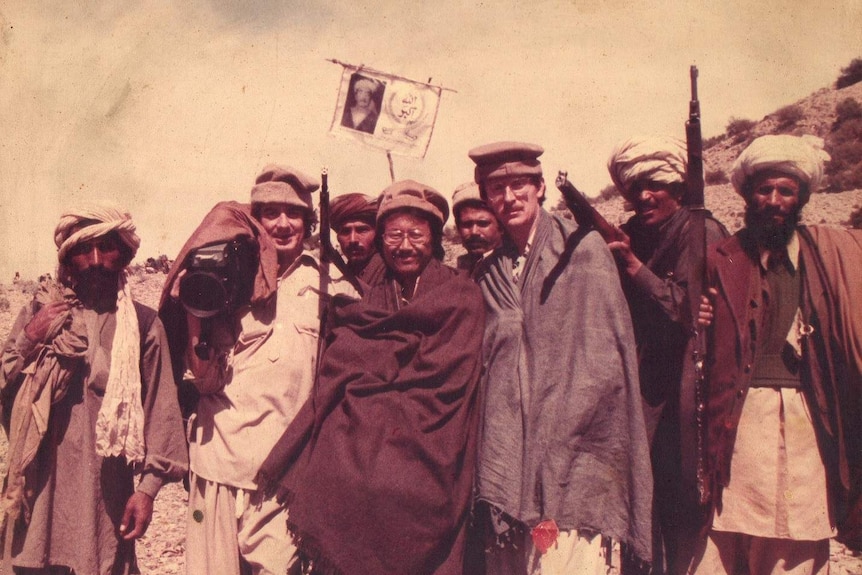 A group of men stand on a mountain in Afghanistan, some holding guns and one man holding a camera.
