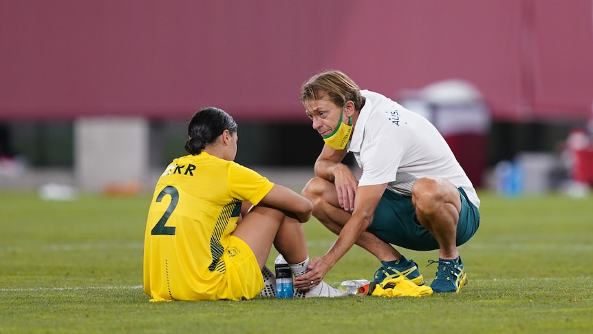 Tony Gustavsson crouches down to console Sam Kerr, who is sitting on the ground and is sad