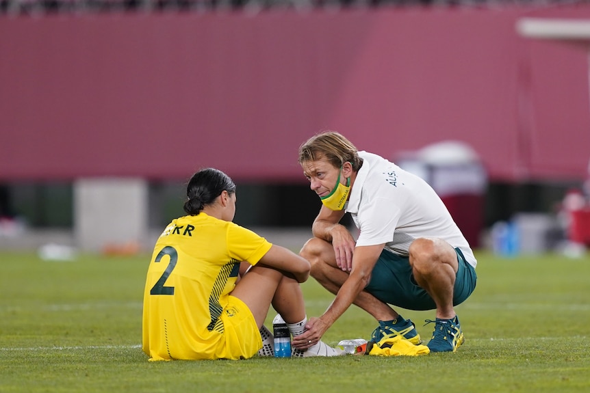 Tony Gustavsson crouches down to console Sam Kerr, who is sitting on the ground and is sad