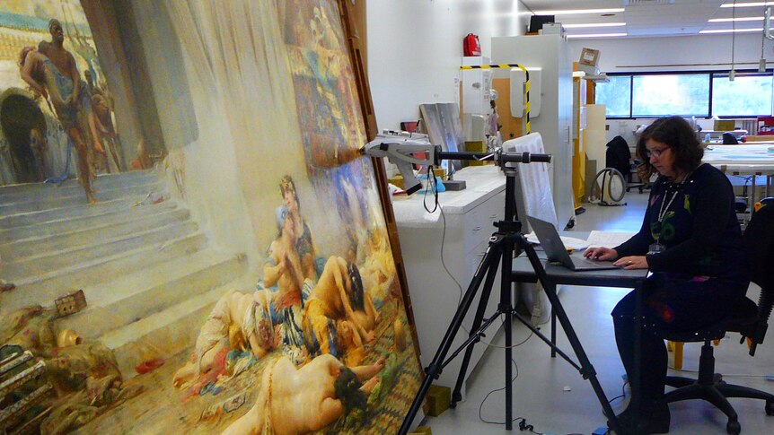 A woman sits at a desk in front of a painting.