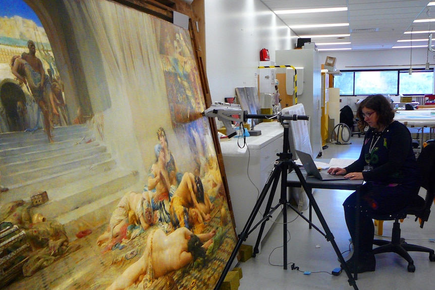 A woman sits at a desk in front of a painting.