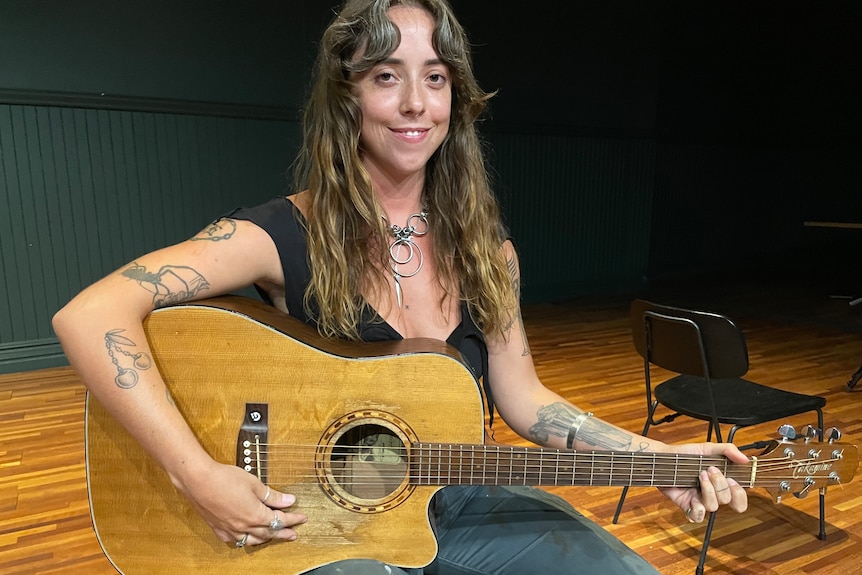 A woman with long hair and tattoos on her arm holds an acoustic guitar. 